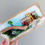 Thumbnail: A piece of wood, painted and decorated with colourful driftwood cottages to create a coastal scene
