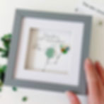 Front view of a hand holding the 17.5cm framed sea glass birthday picture in a grey frame with white mount.