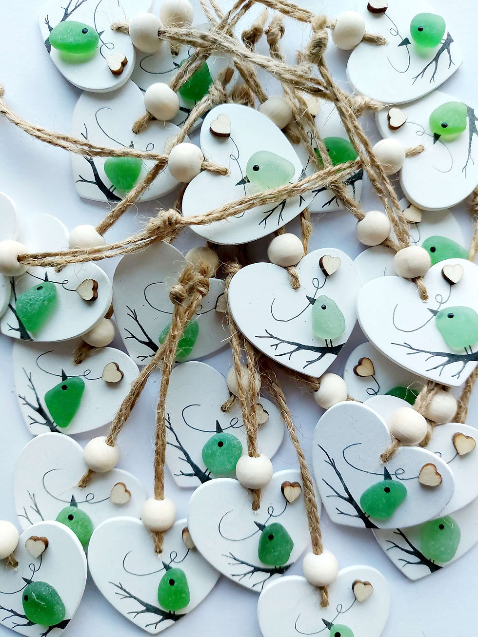 Thumbnail: Pile of white wooden heart hanging decorations decorated with green sea glass birds holding tiny hearts, perched on branches