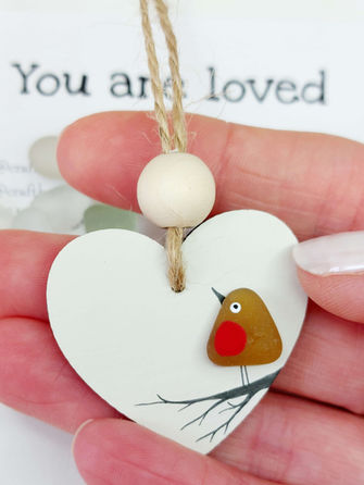 A mini white heart decoration showing a hand drawn branch on which stands a robin made from a brown piece of sea glass.