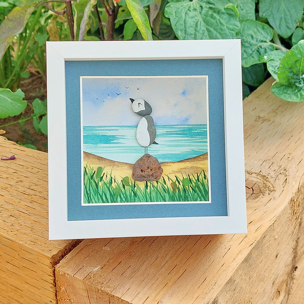 Handmade framed puffin coastal art with painted pebbles, sea glass and a watercolour beach scene in a white frame with blue m