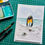 Thumbnail: A black framed 3D watercolour painting of an blue ocean with rocks and ice in the foreground and a pair of pebble penguins.