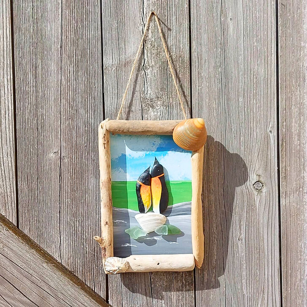 Handmade penguin couple pebble art featuring two hand-painted birds nestled together by the sea, framed with driftwood