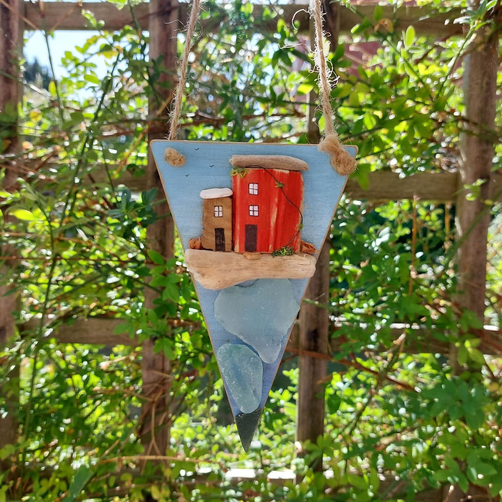 Thumbnail: A pennant shaped seaside decoration. It has been painted blue and decorated with pieces of driftwood houses and sea glass.