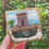 Thumbnail: A square wooden picture, painted blue and decorated with a driftwood cottage and sea glass beach finds.