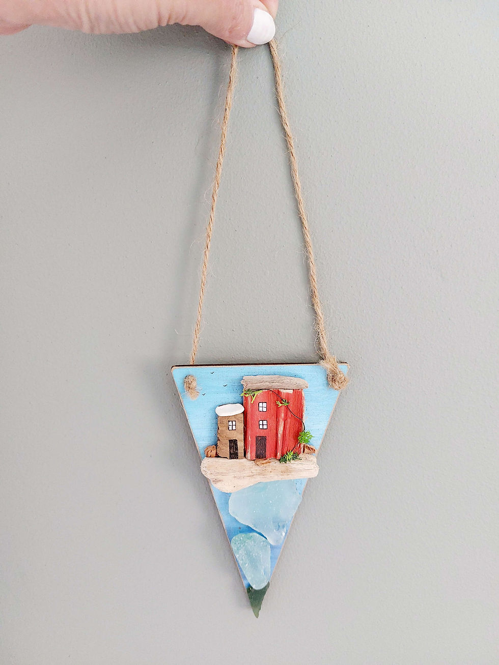 Thumbnail: A pennant shaped seaside decoration. It has been painted blue and decorated with pieces of driftwood houses and sea glass.