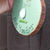 Thumbnail: A hand holding a wooden disc decoration. The wood has been painted green and decorated with sea glass pieces depicting a bird