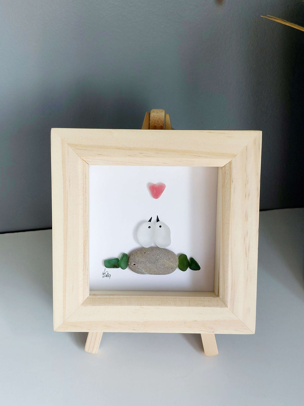 Thumbnail: A pine framed picture of a pair of sea glass 'lovebirds' below a red painted sea glass heart. On a wooden stand.