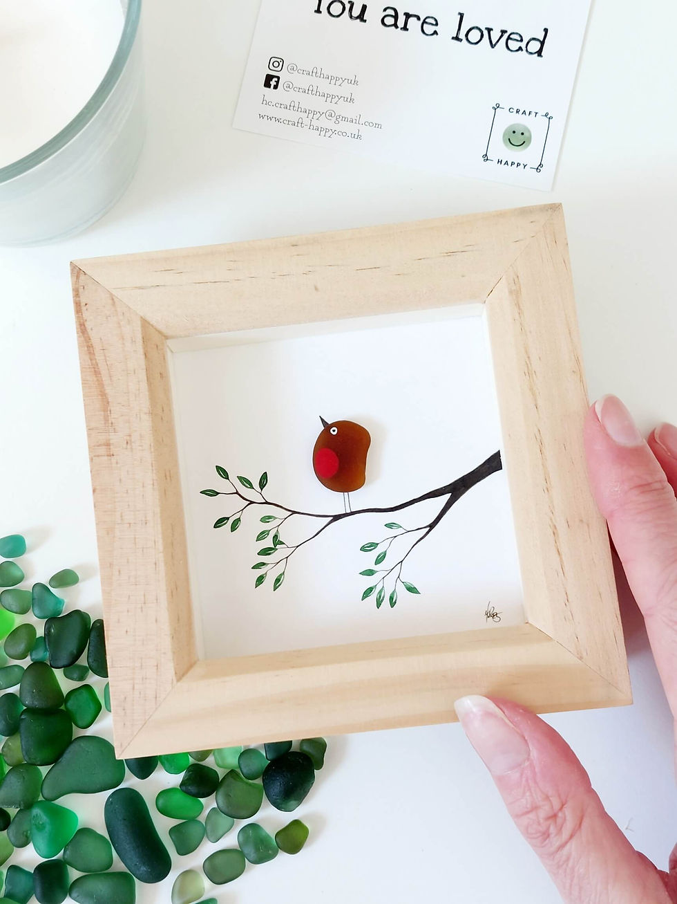 A hand holding a natural pine framed picture of a sea glass robin on a hand painted branch with a twig in its beak.