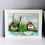 Thumbnail: Framed recycled art consisting of rocky mountains and two alpine cottages on a blue background with a sea glass alpine stream
