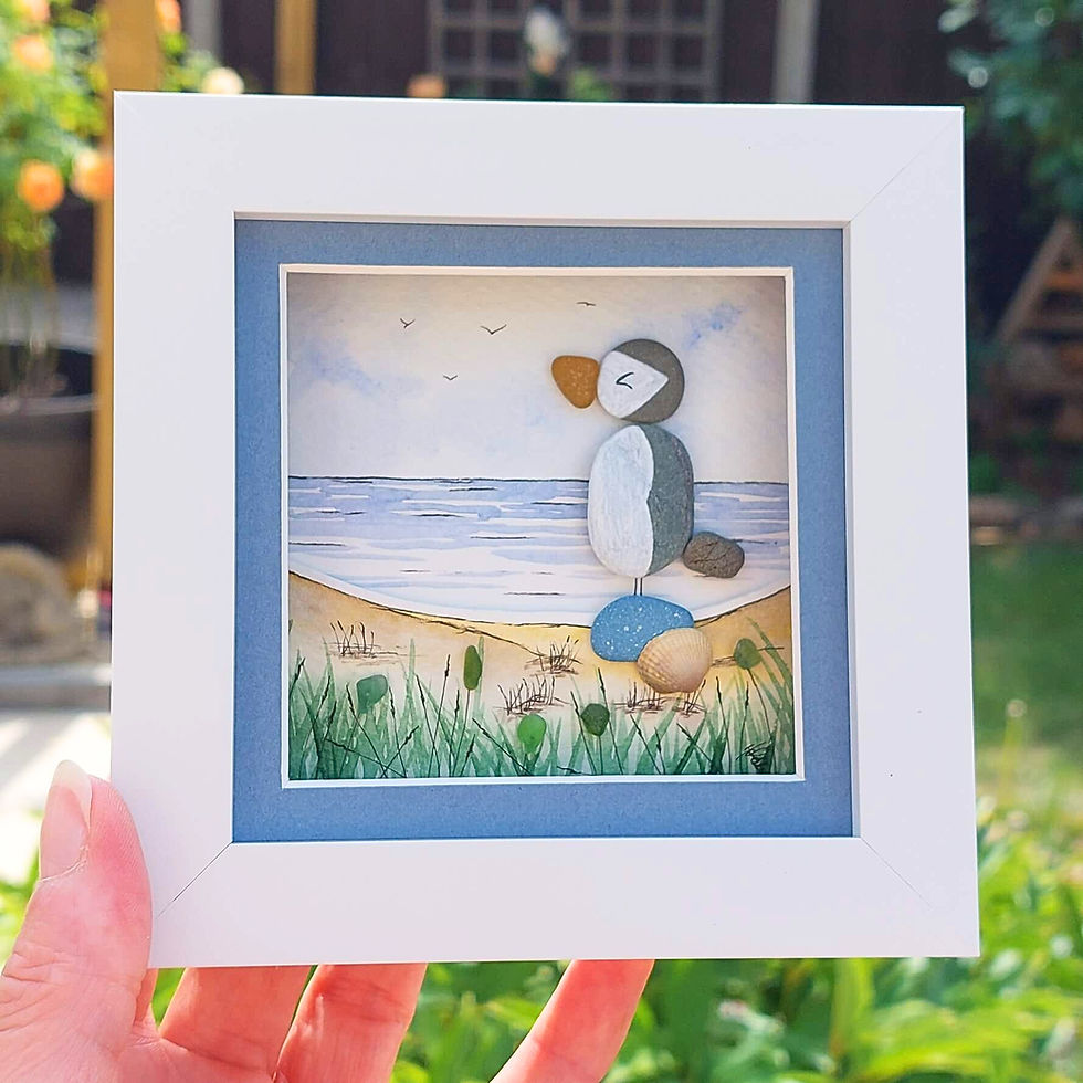 A white framed hand made 3D picture of a puffin made from pebbles with sea glass below on a watercolour background.