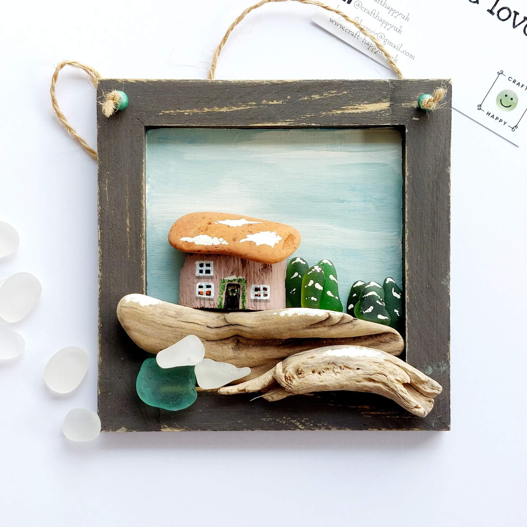 Square wooden wall art in a frame, the outside edge is painted brown and inside decorated with driftwood and sea glass.