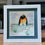 Thumbnail: A white framed hand made 3D watercolour picture showing penguins made from pebbles perched on a piece of sea glass.