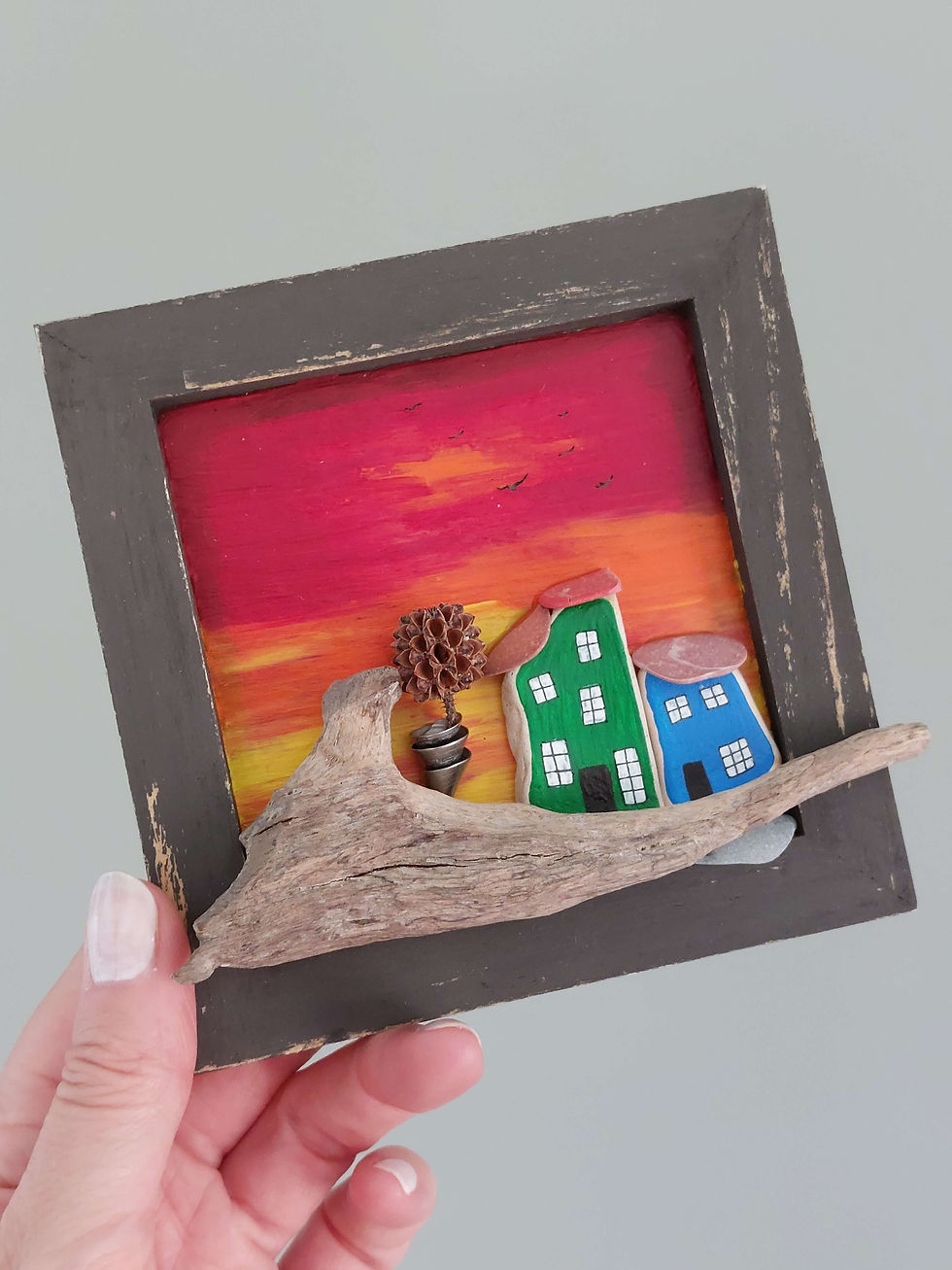 Thumbnail: A hand holding a bright orange and red wooden frame with a coastal cottage scene showing 2 quirky houses on driftwood.