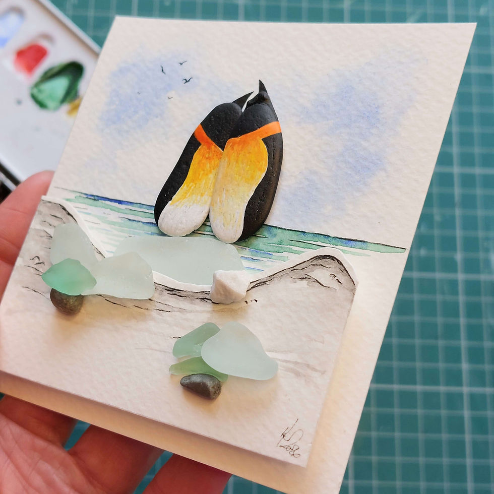 Thumbnail: A white framed hand made 3D watercolour picture showing penguins made from pebbles perched on a piece of sea glass.