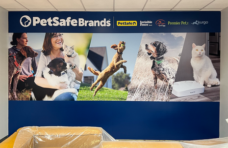 Office wall graphic featuring angled photos of pets and people, with PetSafe Brands and partner logos at the top and a solid blue lower section designed to sit behind furniture in a waiting area.