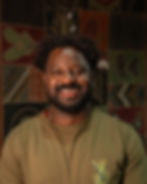 Homem negro sorrindo em retrato, com cabelo crespo e barba, camiseta verde-oliva, fundo com padrões coloridos desfocado.