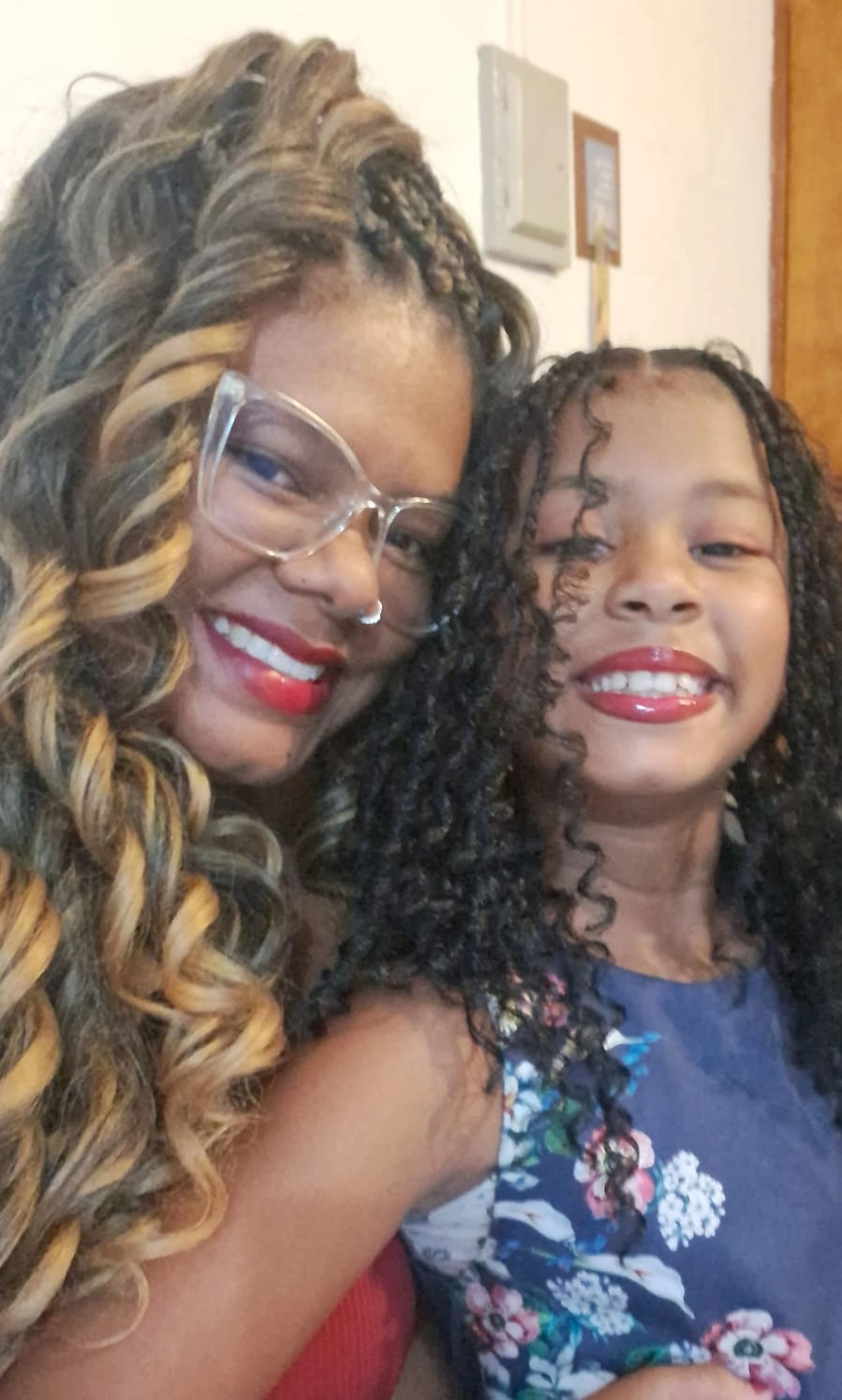 Mulher negra e criança negra sorrindo, com cabelos cacheados. Mulher usa óculos e batom vermelho. Criança veste roupa floral. Fundo neutro. Emocionadas.