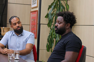 Dois homens conversam em uma sala com parede bege e planta ao fundo. Um usa camisa azul, o outro, camiseta preta. Ambos parecem focados.