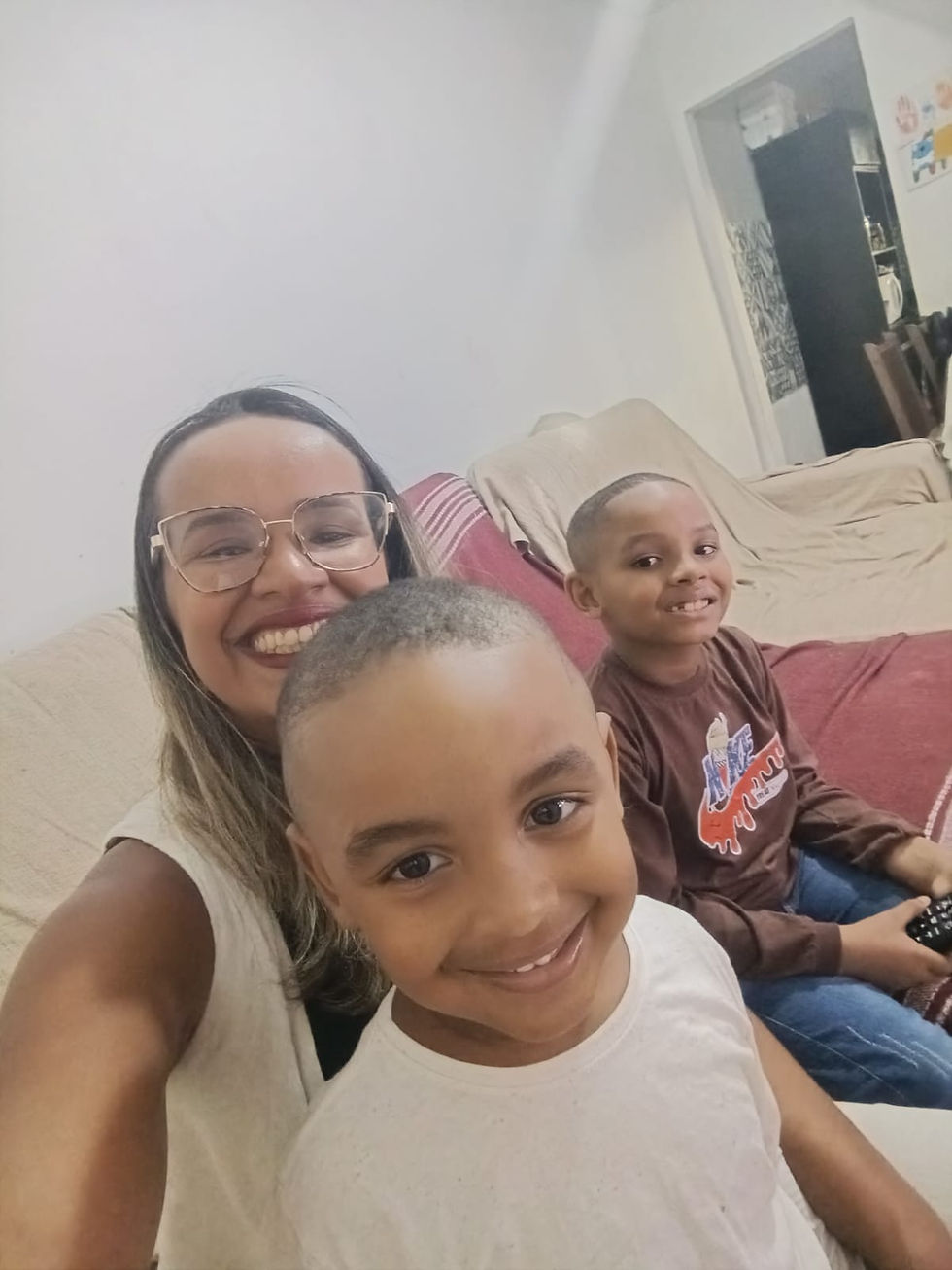 Mulher negras e duas crianças negras sorrindo em um sofá coberto. Uma criança segura um controle remoto. Ambiente acolhedor com cores neutras.