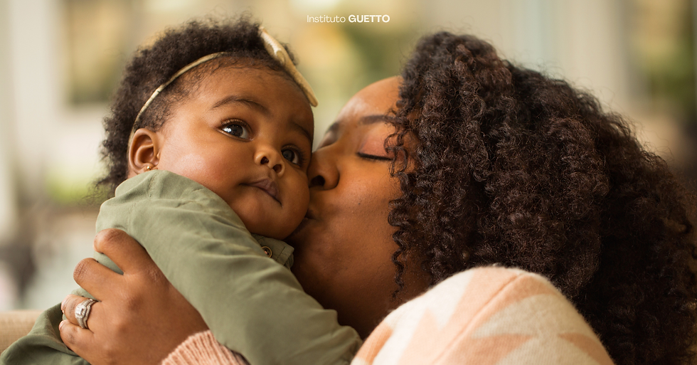 Mulher negra beijando rosto de bebê negro em roupa verde. Cabelo cacheado dos dois. Fundo desfocado. Atmosfera afetuosa.