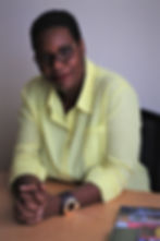 Mulher negra de óculos e camisa amarela, sentada à mesa com as mãos unidas, em ambiente neutro e expressão calma.