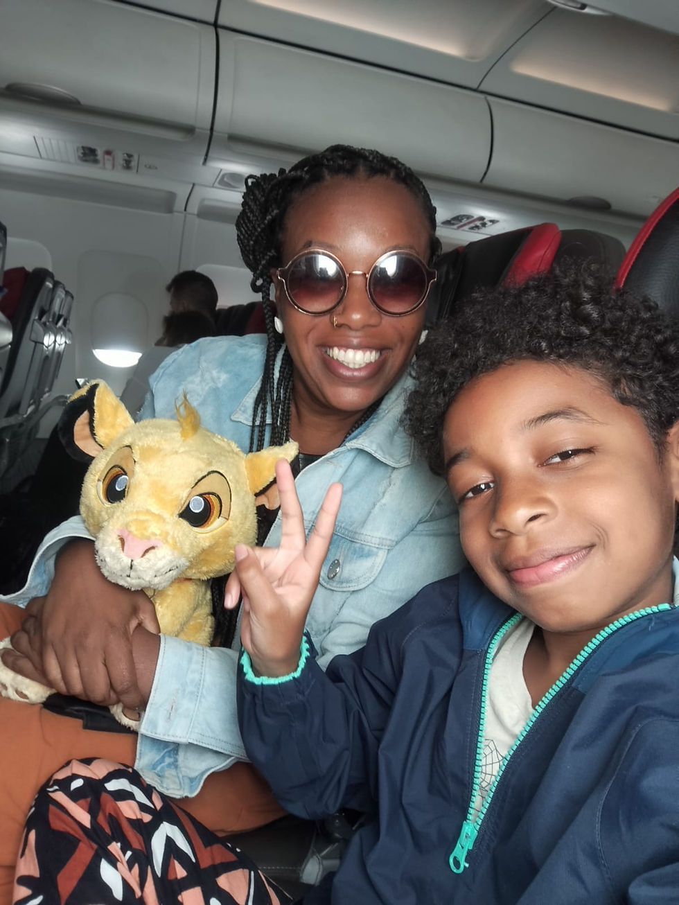 Mulher negra sorridente com óculos escuros e criança fazem gesto de paz em avião. Criança segura pelúcia do Simba. Ambiente alegre e acolhedor.