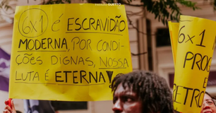 Protesto nas ruas com pessoas segurando cartazes amarelos. Texto no cartaz: "6x1 é escravidão moderna, por condições dignas, nossa luta é eterna".