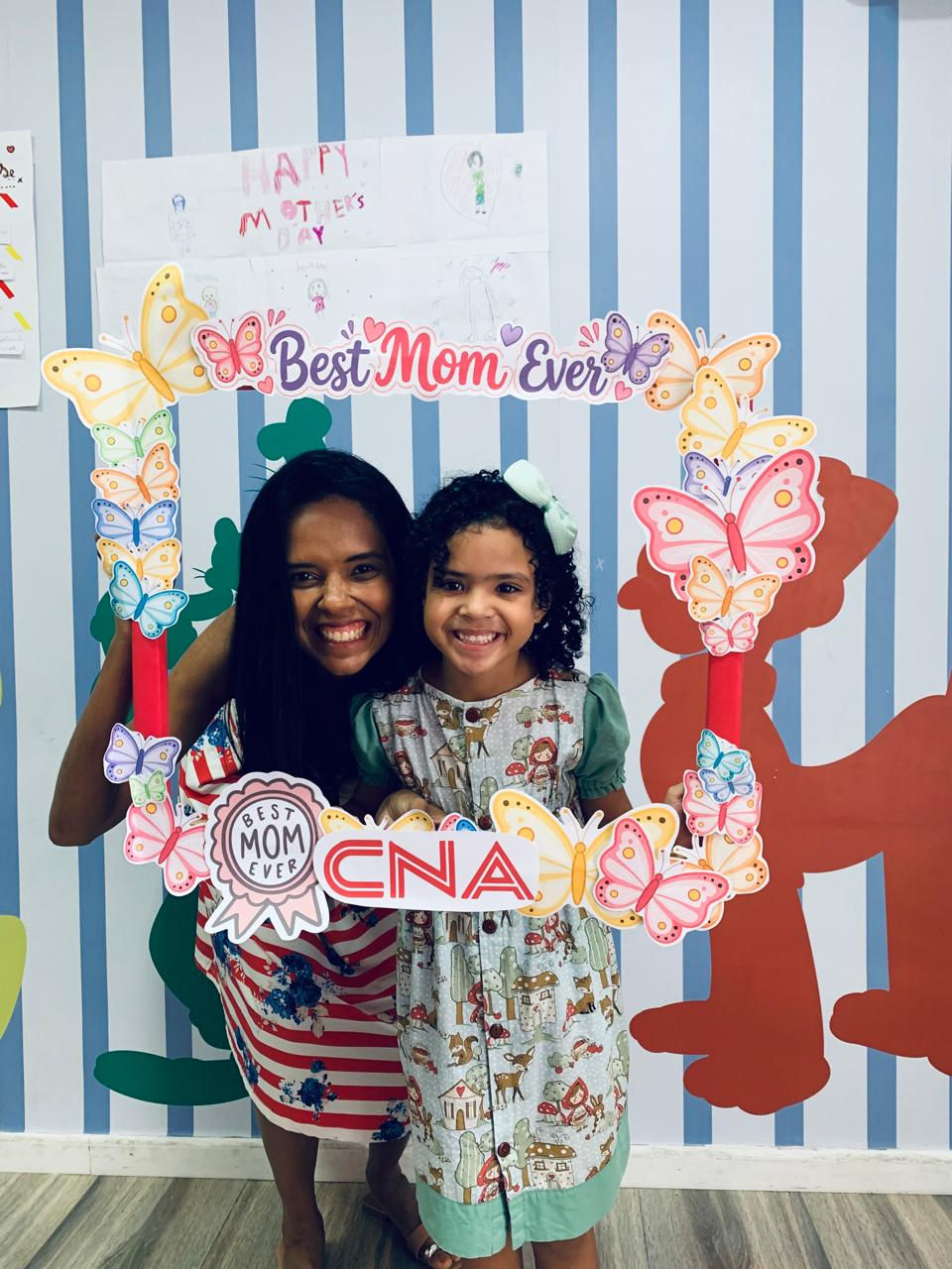 Mãe e filha sorrindo com moldura colorida "Best Mom Ever" e "CNA". Fundo listrado azul e branco, desenhos de borboletas e mural. Em clima de Dia das Mães. Ambas são negras.