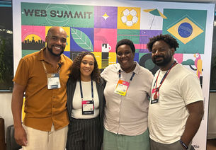 Quatro pessoas negras sorridentes posam no Web Summit diante de painel colorido com ícones e crachás.