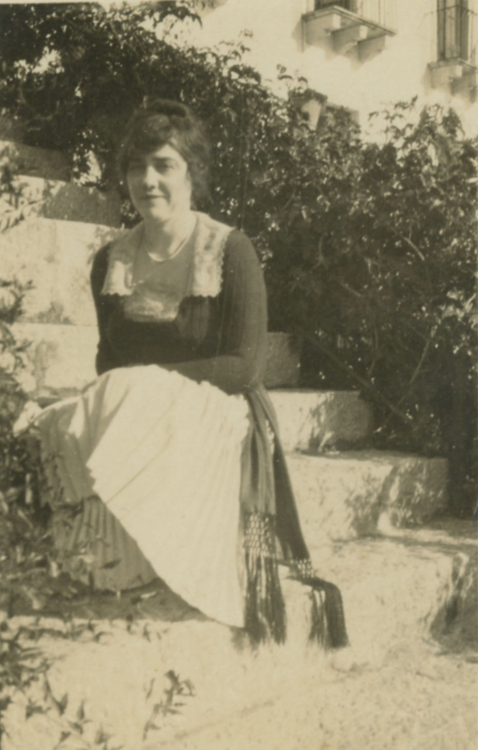 A sepia tone photograph of Nell C. Milligan sitting outside on a staircase flanked by shrubs.