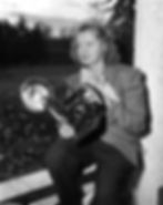 Betty Love poses with her camera. Love is seated on a railing; a yard is in the background. She holds a camera with a large flash bulb.