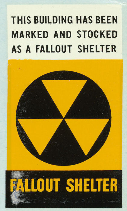 A decal used to mark community fallout shelters. The Civil Defense's fallout shelter signal (three yellow triangles in a black circle) occupies the vertical center of the sign. Text at top reads: "This building has been marked and stocked as a fallout shelter." Text at bottom reads: "Fallout shelter."