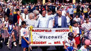 President George H. W. Bush and First Lady Barbara Bush lead the Fourth of July parade in Marshfield, 1991. Many people fill the frame. A sign
