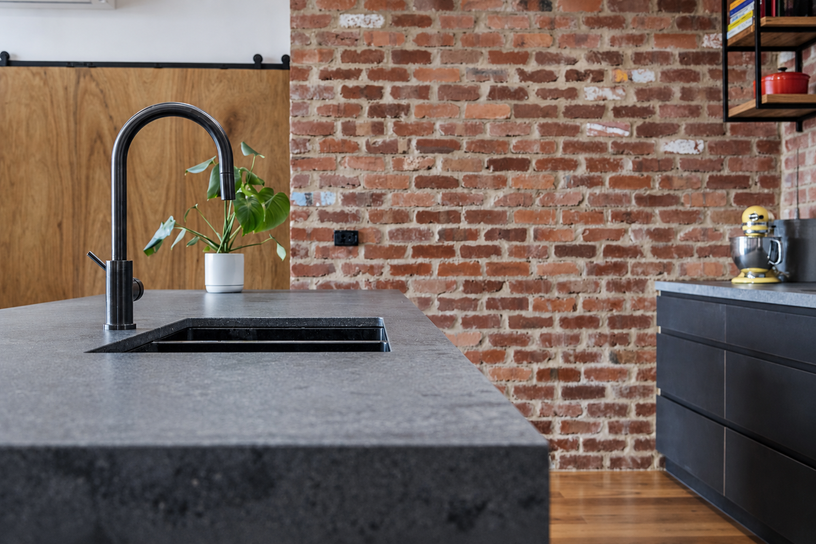 Kitchen island detail with dark tapware, grey benchtop and recycled brick forming a textured backdrop.