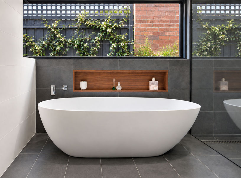 Freestanding white bath against a dark grey tiled wall, with a timber-lined niche and a wide window overlooking greenery and brickwork.