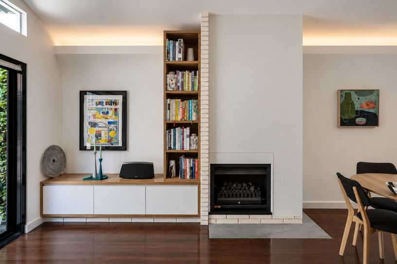 Living room joinery with low white cabinetry, a tall bookshelf and an adjoining fireplace beneath softly lit ceiling recesses.