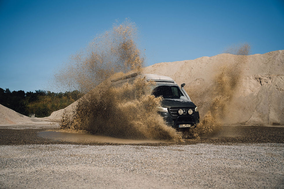 Geländewagen spritzt in einer Kiesgrube Wasser und Sand auf, vor blauem Himmel und hellen Sandhügeln.