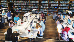 People studying in a library with computers and books. Wooden floor, bookshelves in background. Focused atmosphere, diverse group.