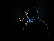 Pessoa em ambiente escuro usando moletom com capuz e máscara neon azul com desenho de olhos em “X” e sorriso estilizado, iluminada contra um fundo noturno.
