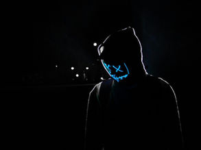 Pessoa em ambiente escuro usando moletom com capuz e máscara neon azul com desenho de olhos em “X” e sorriso estilizado, iluminada contra um fundo noturno.