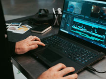 Pessoa usando um laptop para editar um vídeo em uma linha do tempo de edição, sobre uma mesa com fotos impressas e um caderno ao lado.