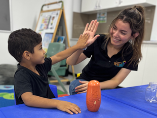 Boy and educator high-five and smile at each other. They are sitting at a blue table in a classroom setting.