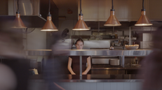 Still of lead actress, Casey, standing behind a kitchen counter, various copper lamps above her. Slow shutter speed shot so people are blurry in front of her. 