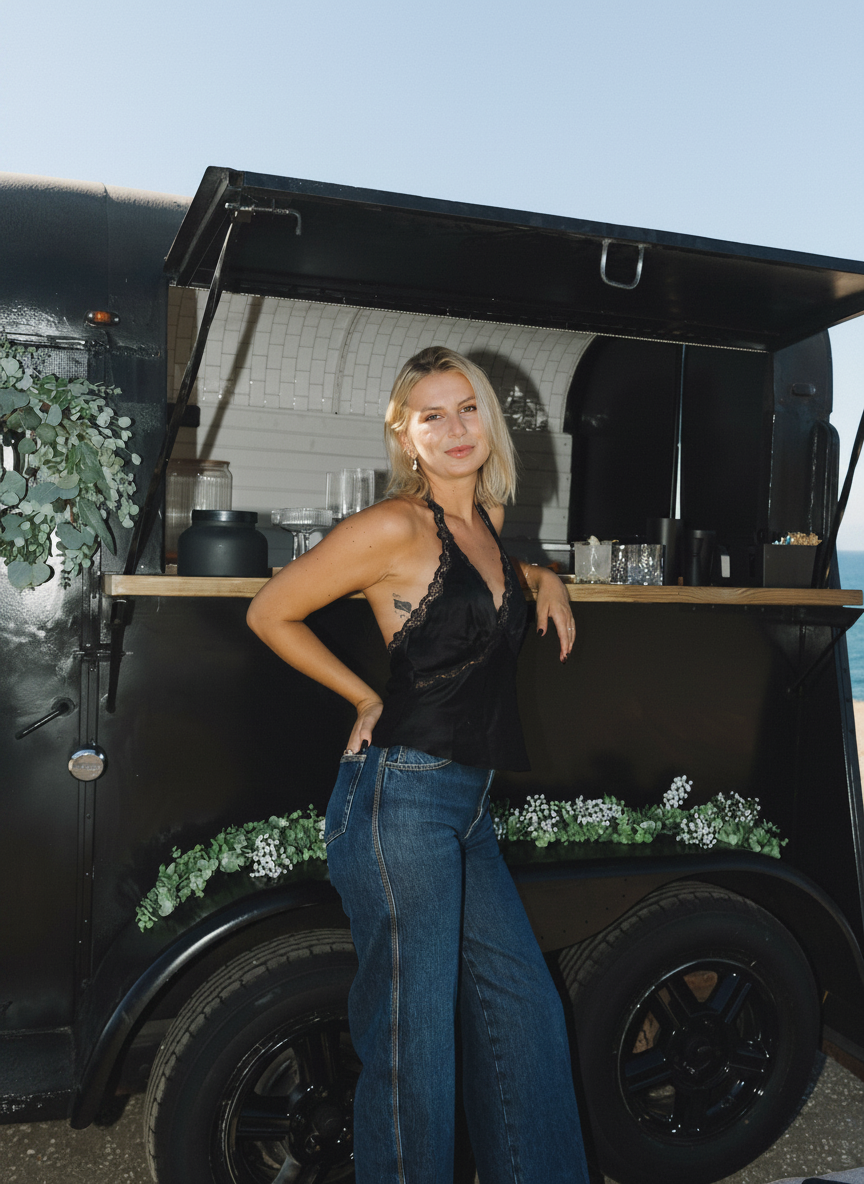 Portrait of a woman leaning against a black mobile bar trailer with ocean in the background, vertical composition.