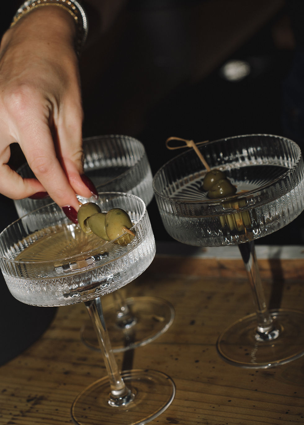 Close-up of a hand placing olives into elegant coupe glasses on a wooden bar, warm moody lighting