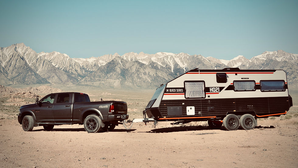 Camioneta gris con tráiler "Black Series" en paisaje montañoso. Cielo azul, montañas nevadas al fondo. Ambiente de aventura.