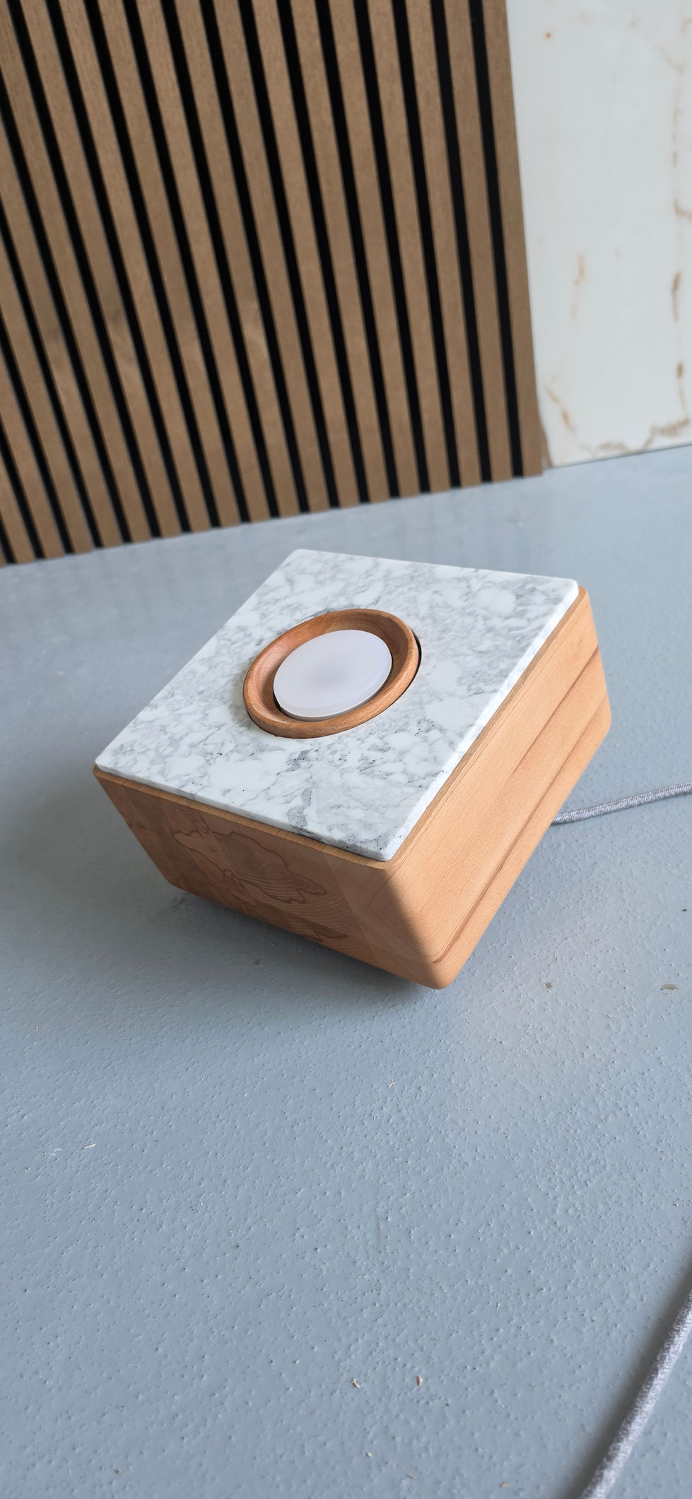 Square floor spotlight made of heartwood beech wood and white-grey marbled natural stone with a central round light source; the spotlight stands on a structured grey floor, next to it wooden slats and a brown-white grained stone surface.