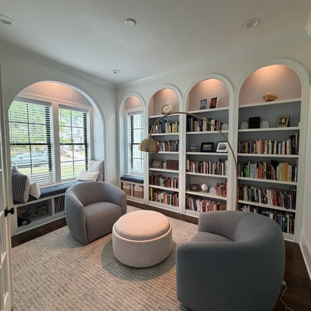 Custom arched built-in bookshelves with down lighting, blue painted interiors, white face frames, and a window bench nook in a styled Raleigh home library.