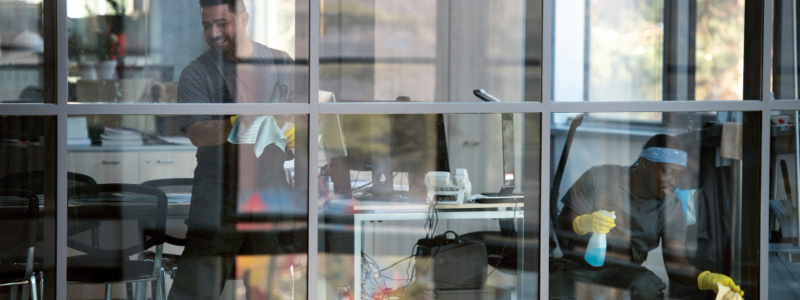 Two men clean a glass-walled office, one wiping and one spraying cleaner, with desks and chairs in the background.